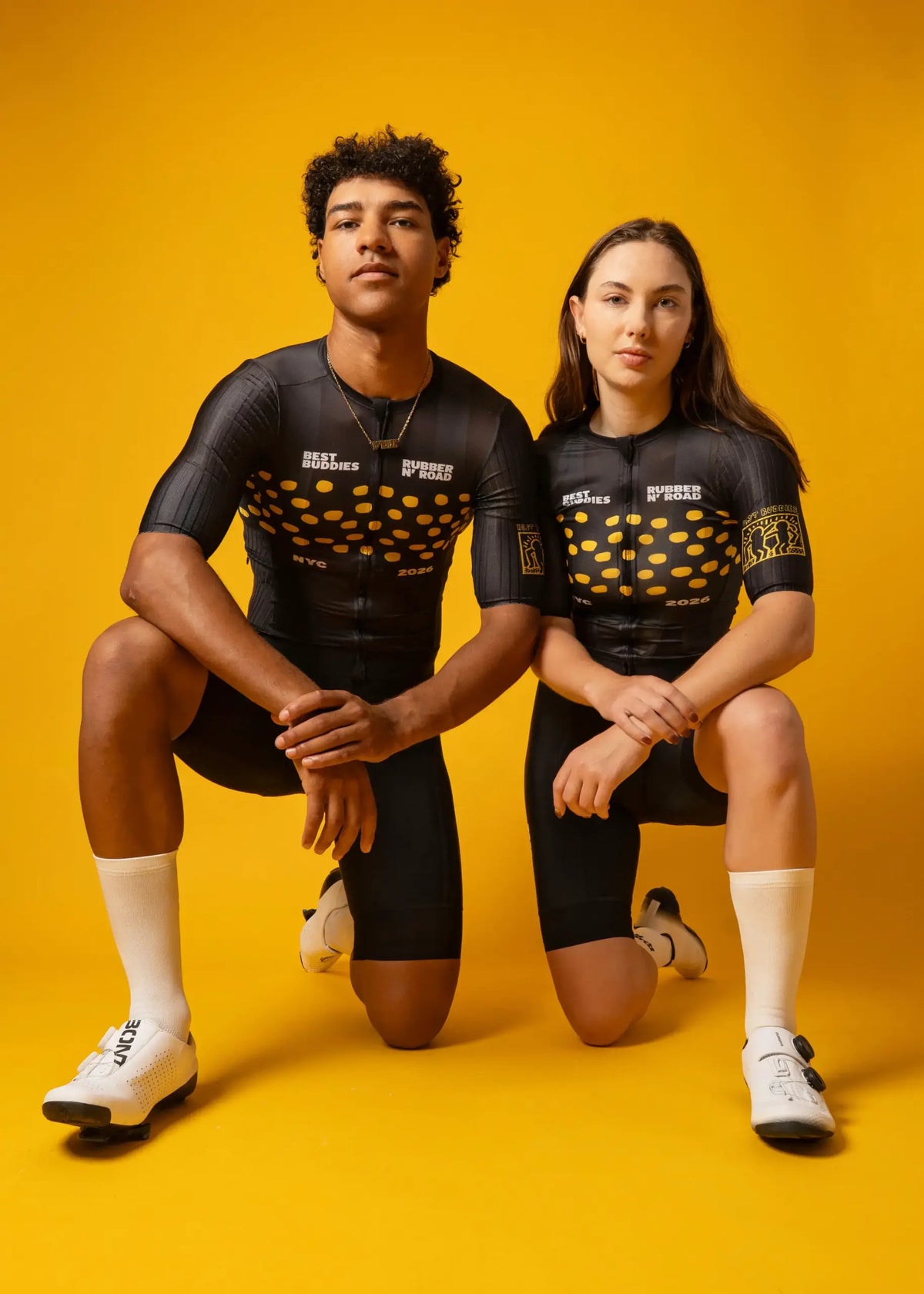 Two cyclists posing in matching black and gold cycling jerseys and shorts against a solid yellow background.