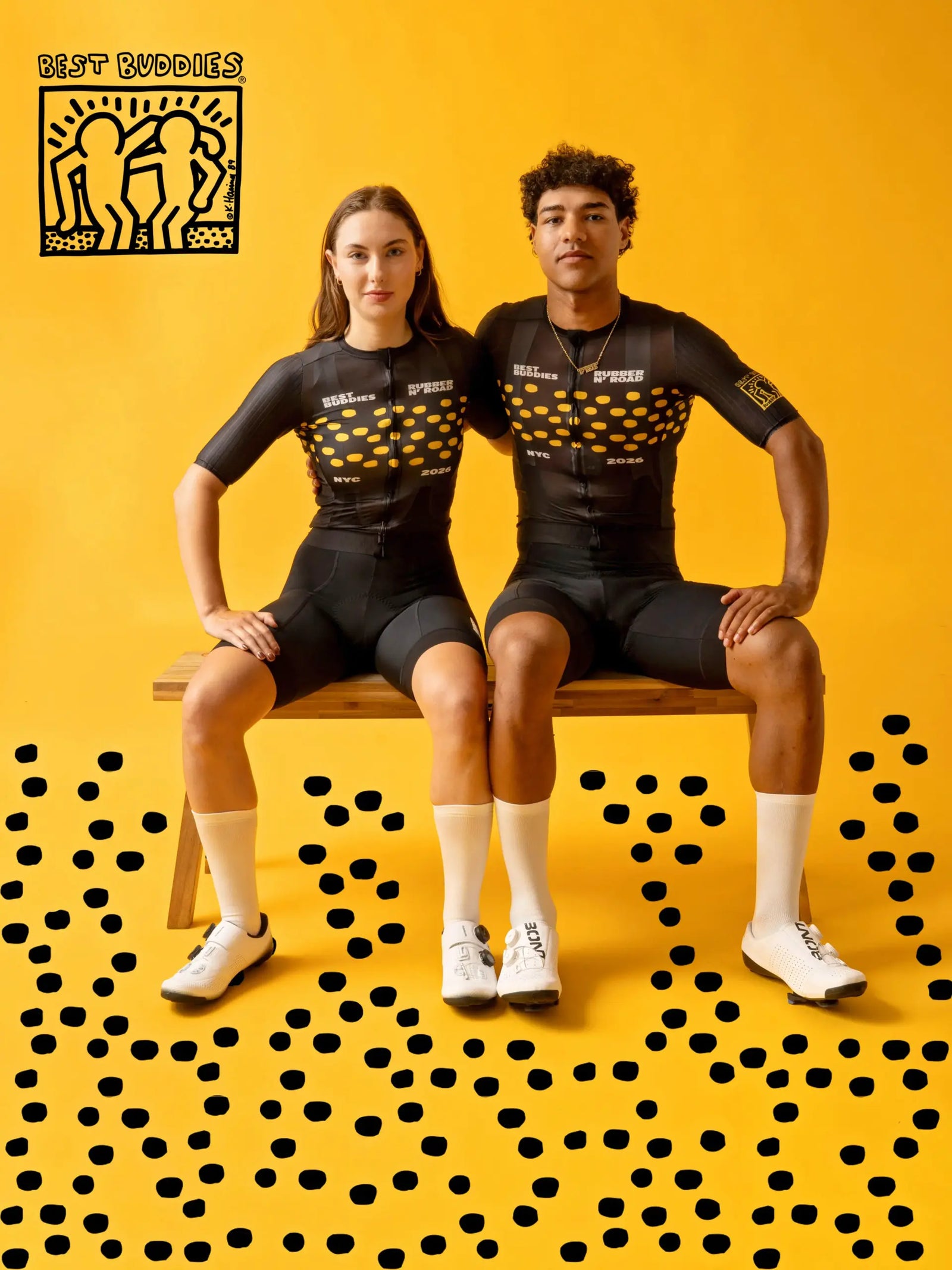 Two cyclists sitting on a wooden bench wearing matching black and yellow Keith Haring cycling jerseys and shorts, posed against a bright yellow background with black polka dot patterns.