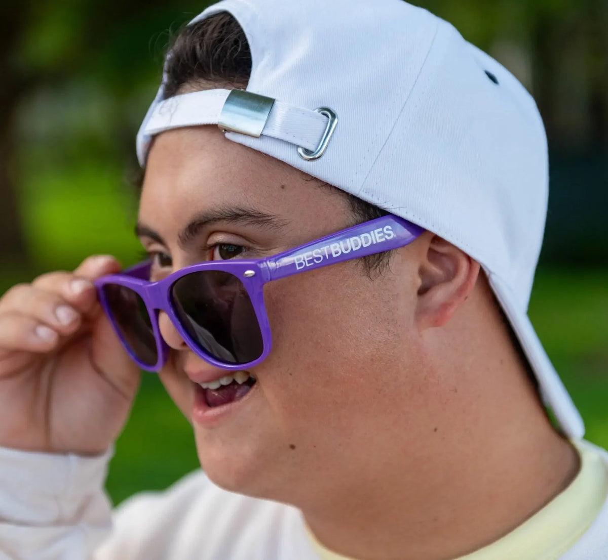 A young man wearing a white baseball cap backward and a light-colored long-sleeve shirt adjusts his purple sunglasses with "BEST BUDDIES" printed on the side. He is smiling and looking off to the side, with a blurred green outdoor background.